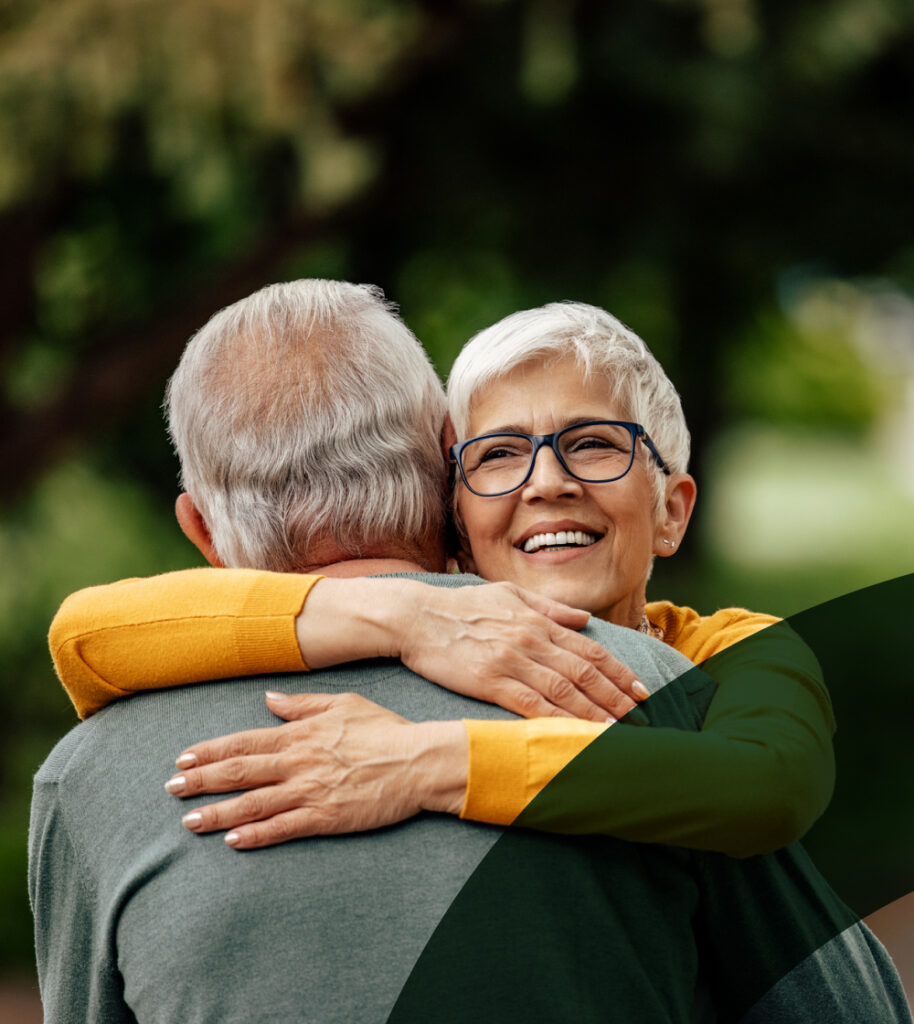 Two people with grey hair embracing warmly in a lush green outdoor setting.