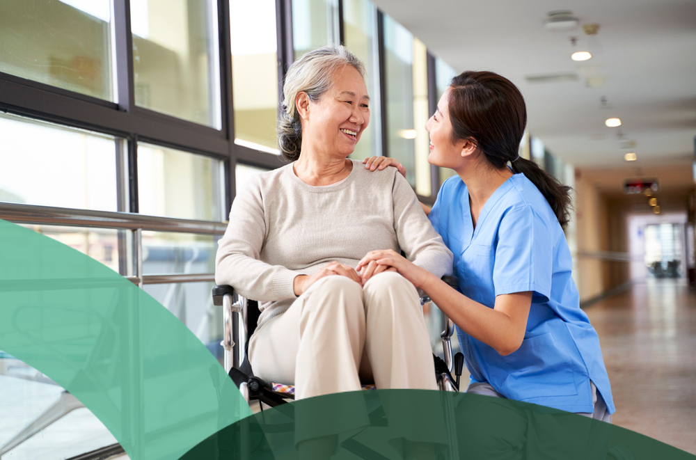 Healthcare worker smiling at a woman in a wheelchair in a bright hallway.