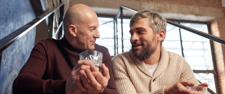 Two men sit on stairs having a heartfelt conversation over drinks.
