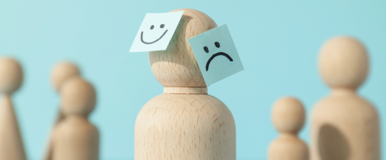 Wooden figure with happy and sad face paper notes on light blue background.